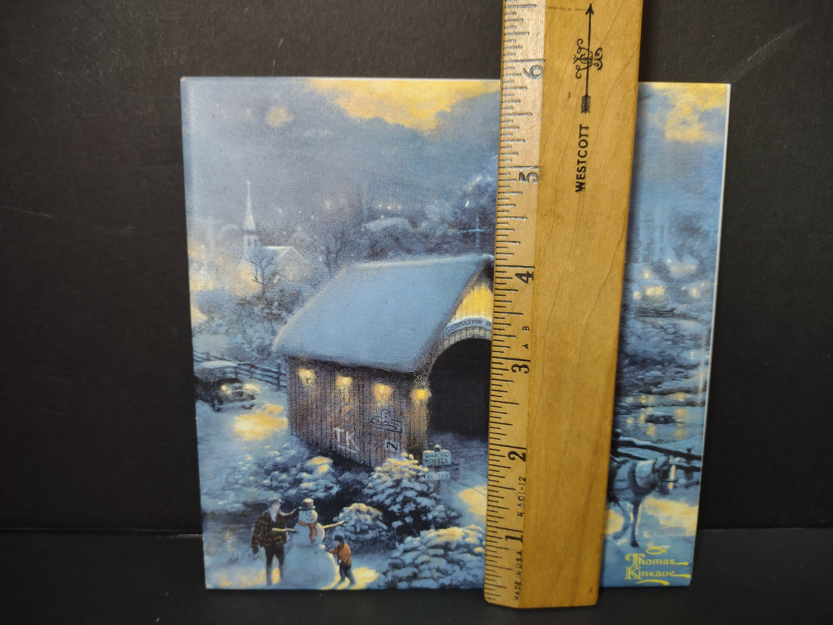 Thomas Kinkade Winter Evening Memories ceramic tile showing snowy winter village with covered bridge, snowman, and warm glowing light, with ruler showing approximately 6 inch size