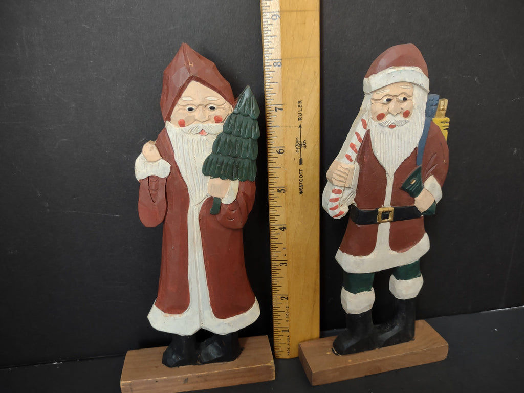 Two vintage folk art painted wood Santa figurines on wooden bases, approximately 8 inches tall — left Santa in long red robe holding Christmas tree, right Santa in red coat holding candy cane with toy sack on back