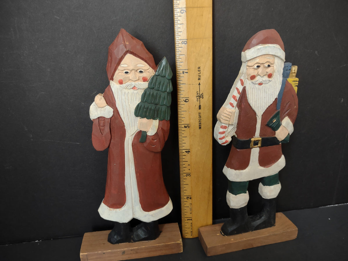 Two vintage folk art painted wood Santa figurines on wooden bases, approximately 8 inches tall — left Santa in long red robe holding Christmas tree, right Santa in red coat holding candy cane with toy sack on back