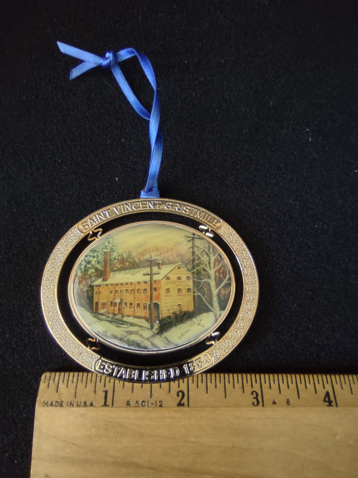 Saint Vincent Gristmill ornament with gold-tone frame winter scene and blue ribbon beside ruler